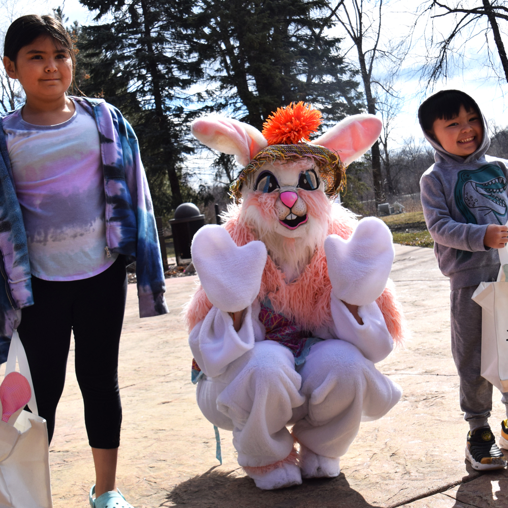 Person dressed as an Easter bunny with two young children on either side outdoors at city hall