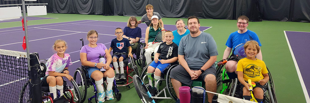 Four kids and two adults in wheelchairs holding tennis rackets in front of net with a woman in red standing behind them